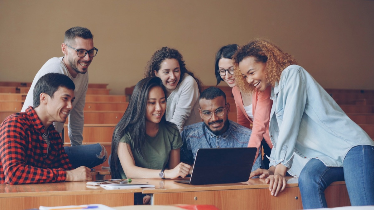 A group of students leaning over a laptop together in a lecture hall, smiling